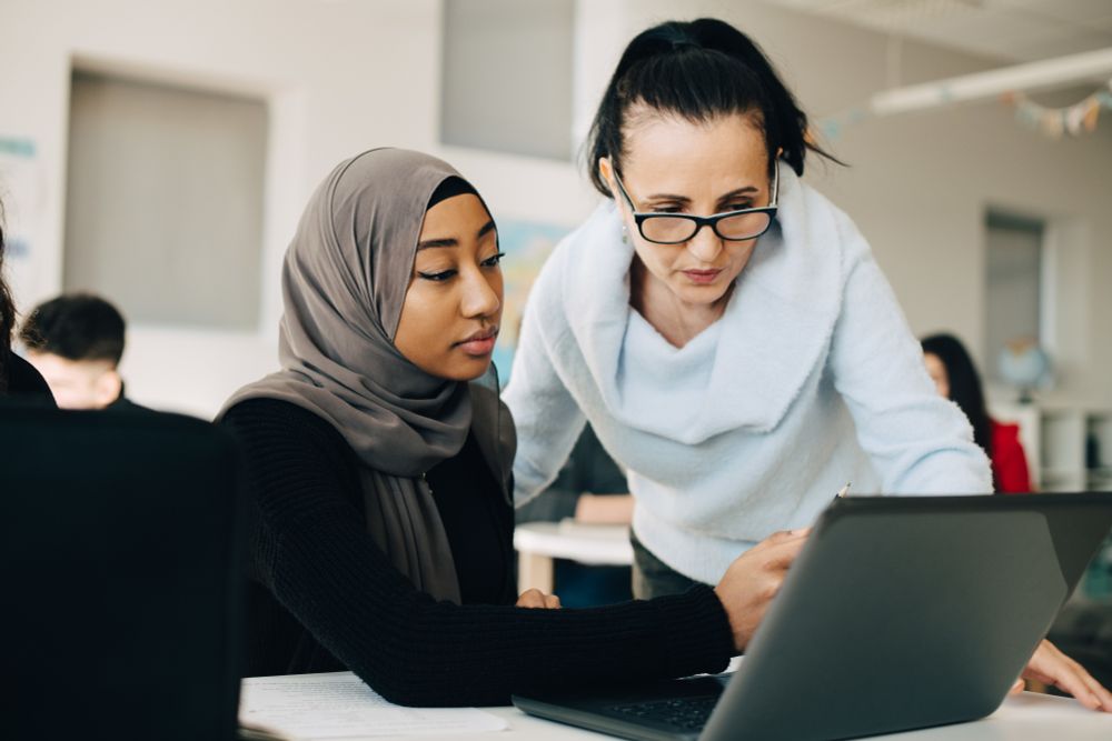 A woman is standing by the desk of a younger woman in a training room and appears to be helping her.