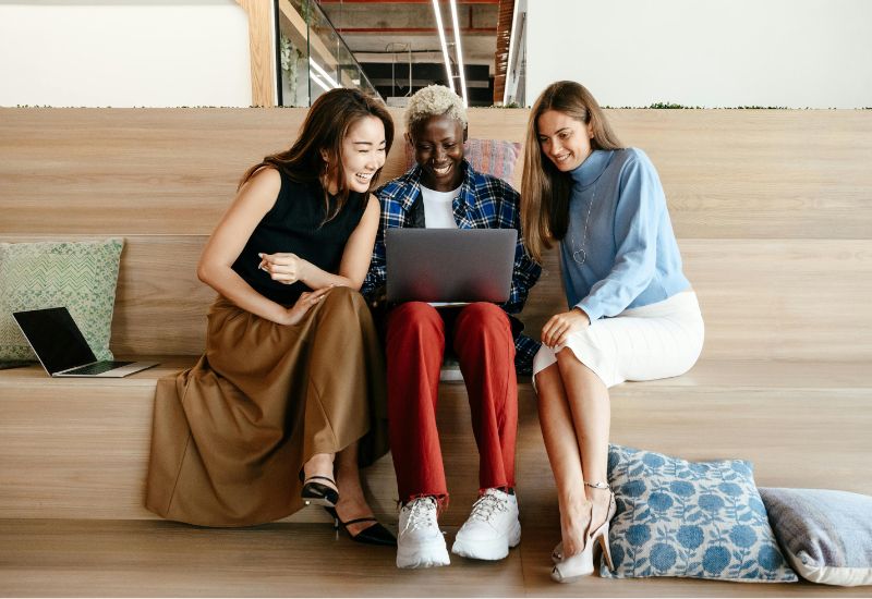 Three women are sitting on a bench gathered round one laptop, they are smiling and looking at the screen