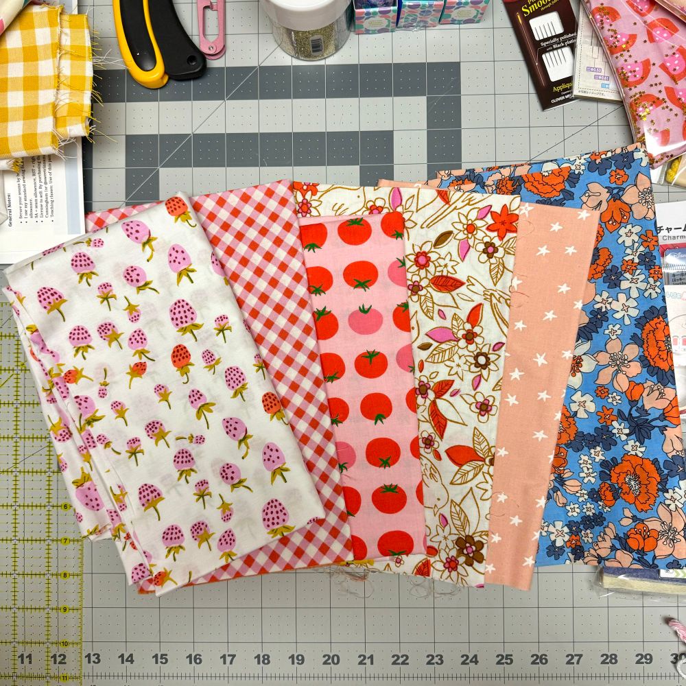 Six fabrics in various shades of pink, red, peach and blue are spread out on a cutting mat. 