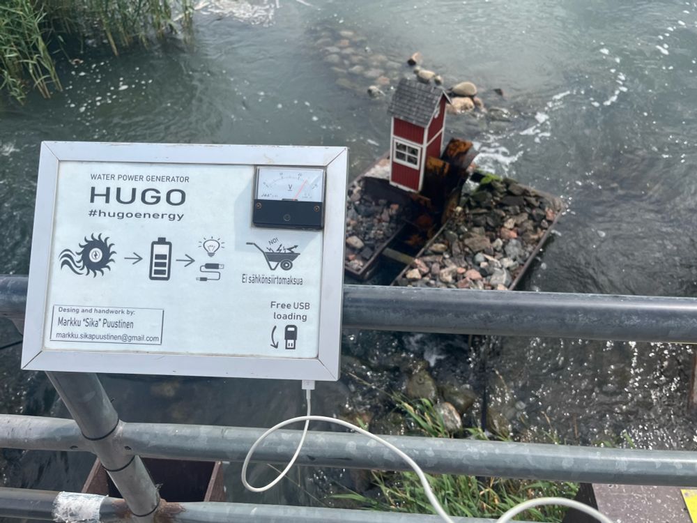 There is a sign saying “water power generator HUGO #hugoEnergy” then a wind turbine symbol with an arrow to an empty battery symbol then an arrow to a cable. It also has “free usb loading” with an arrow and a usb iPhone charging cable coming out of it. Behind the sign in running water there is a mini water turbine with a mini house attached to it. The turbine is supplying energy to the charger.