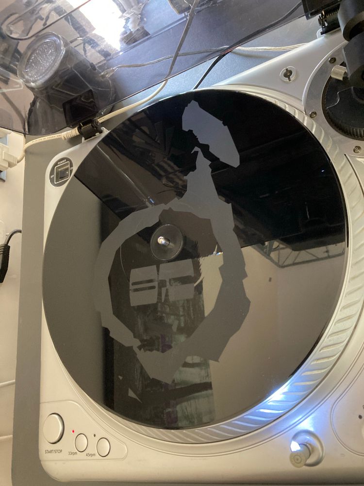 A white label (test pressing of a record), sits on a record deck. The back of the record is very shiny with a logo in mat- something I have never seen before.