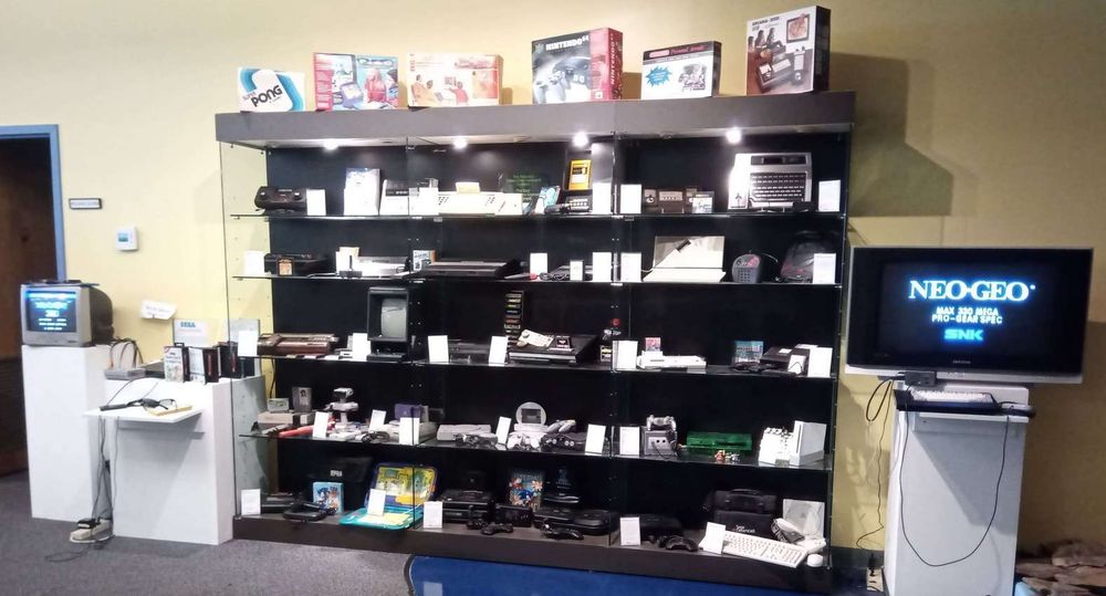 A variety of 30 game systems and ephemera in a display case, with six boxes on top. A Sega Master System is running Zaxxon 3D on the left, and a Neo Geo is booting on the right-hand TV.
