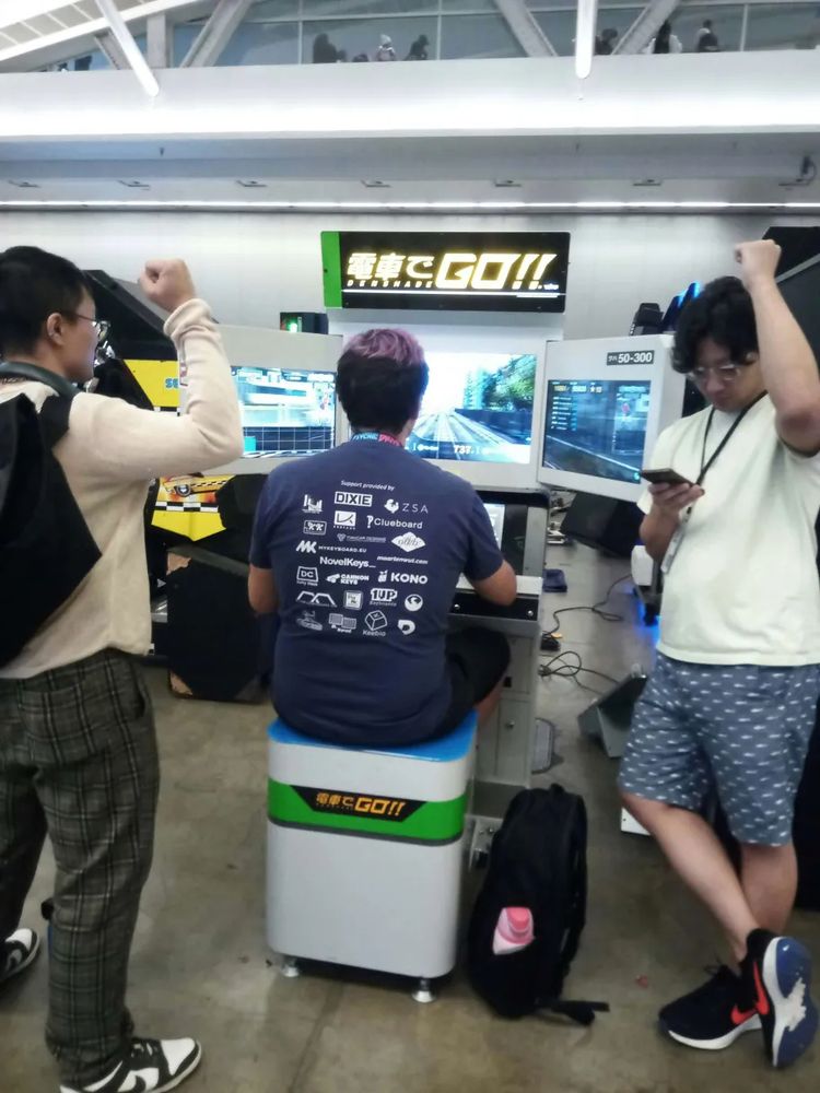 three dudes in front of a train simulator arcade game - densha de go!!