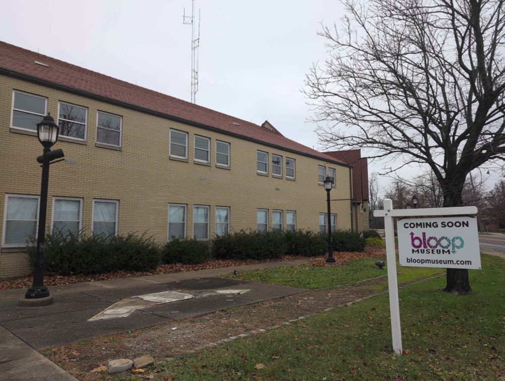 A view of a sidewalk in front of an old beige brick municipal building with a red roof, and "coming soon: bloop museum" on a signpost out front. The scenery includes a tree that has lost its leaves from fall near the sign, and a tall radio tower behind the building.