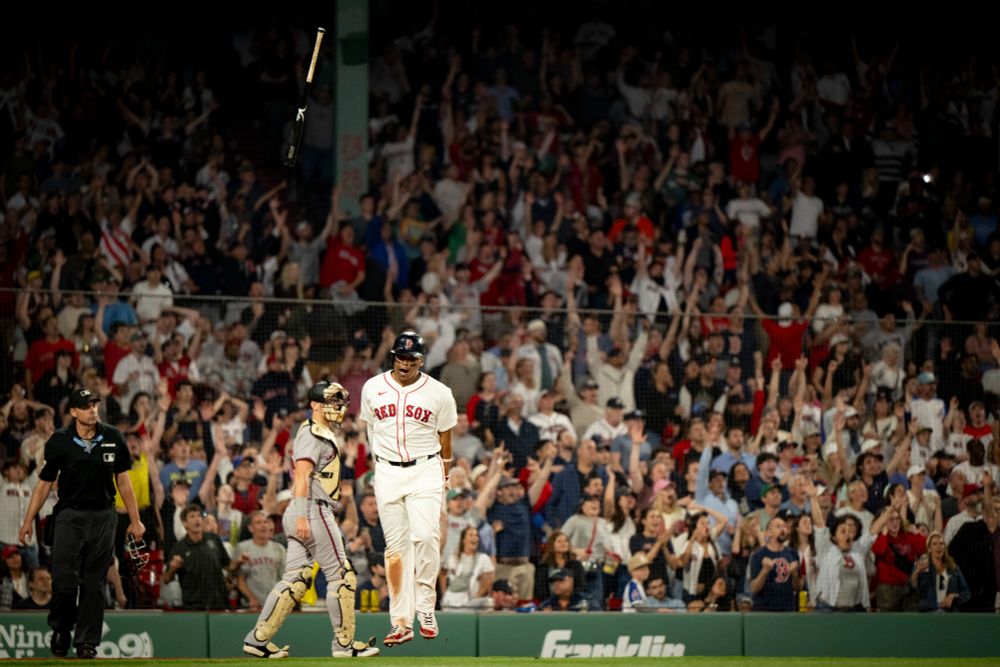 Rafael Devers jumps and shouts in celebration as he turns down the first base line and flips his bat several feet above his head at Fenway Park. The Fenway crowd stands and cheers behind him. 