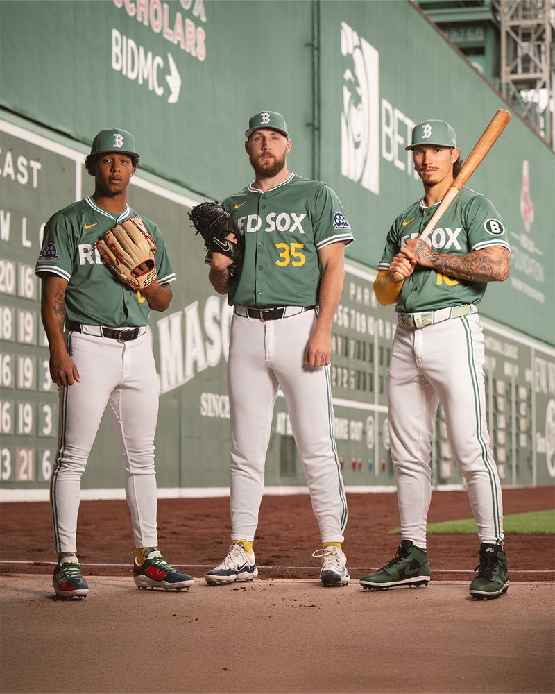 Brayan Bello, Garrett Crochet, and Jarren Duran all stand together posing for a group portrait down the left field line at Fenway Park with the Green Monster in the background. All three wear the Red Sox City Connect uniforms with green jerseys the same shade as the Green Monster, reading “RED SOX” across the front in white with their respective numbers in yellow. Green hats display the Red Sox “B” logo in white, and pants are white with green piping detail downs the sides. 