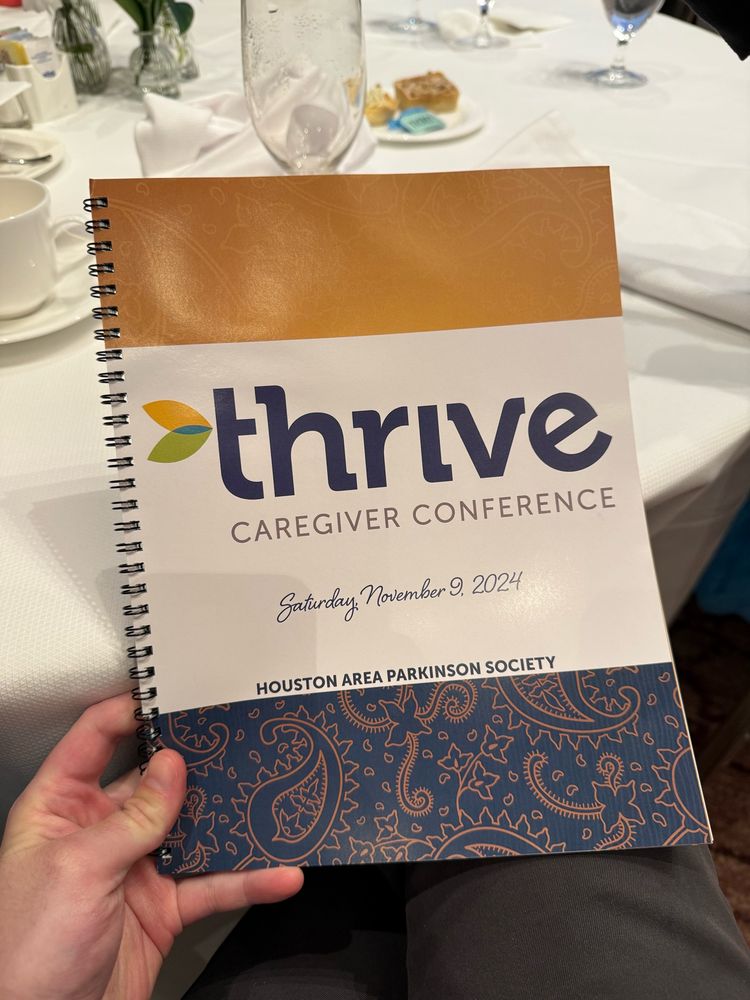 A close-up image of a spiral-bound program booklet for the “Thrive Caregiver Conference” held on Saturday, November 9, 2024, organized by the Houston Area Parkinson Society. The cover features a blue and gold paisley design, adding a decorative element beneath the event details.