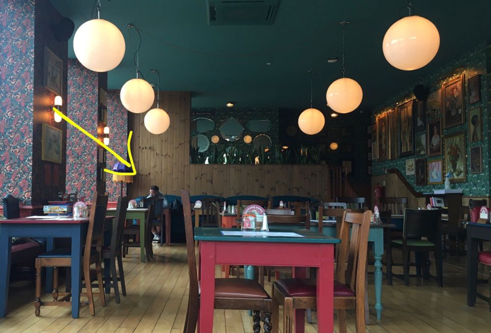 The location is the interior of a quirky cafe. High ceilings, large globe lamps hanging from the ceiling, the walls crammed with framed paintings (over strongly patterned wallpaper). 
Left of centre, at the far end of the room from where I’m sitting, is a man at a table. He is marked with a large yellow arrow!