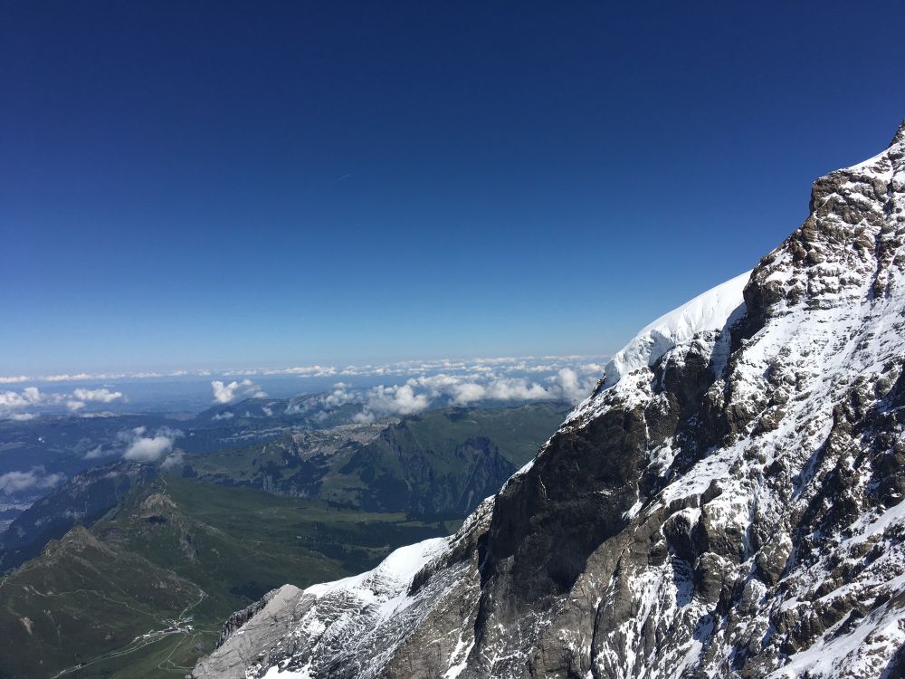 Looking out over northern Switzerland. Green mountains and scattered cumulus clouds (seen from above, n the left, and the ragged, snow-splattered slope of the Mõnch to the right. Deep blue sky above, fading to pale blue of the horizon. 