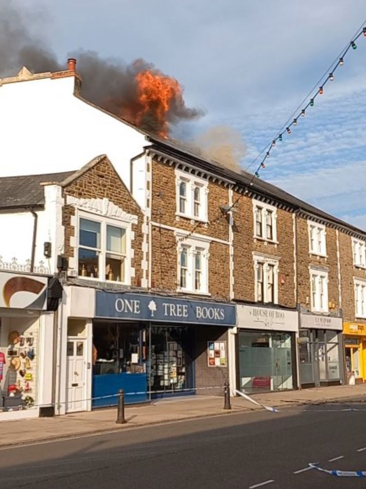 The image shows an angled view of a row of four shops (stretching from the left of the image and away to the right), taken from across the road. The three storey terrace is built of stone, with a white-painted gable end (left). Flames, which started in the property next door, have spread to the roof of One Tree Books (the left-most frontage just before centre), and can clearly be seen against the cloudy/blue sky above. In the end, the whole block (at least six businesses and a number of residential flats) were gutted. 