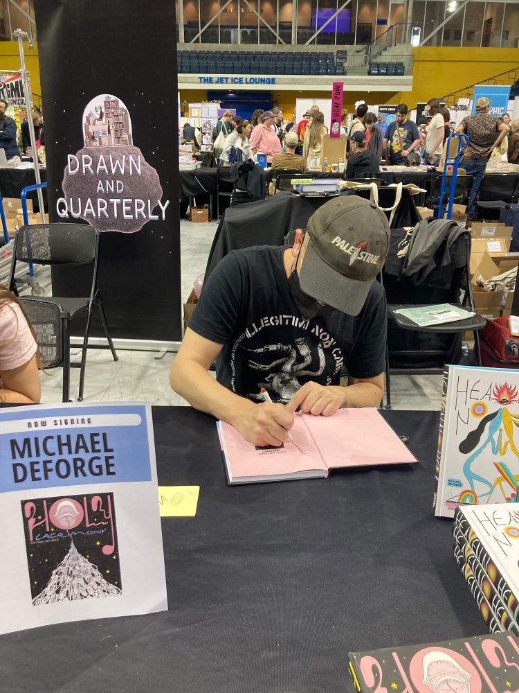 photo of Michael DeForge, sitting at a table signing their book Holy Lacrimony for me. 