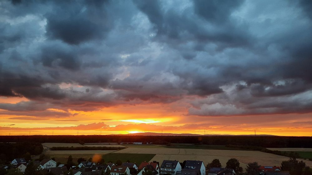 dramatische, blaugraue Gewitterwolken, sie werden von unten von der untergehenden Sonne am Horizont in orange beschienen. Darunter sieht man ein abgeerntetes Getreidefeld und einige kleine Einfamilienhäuser.