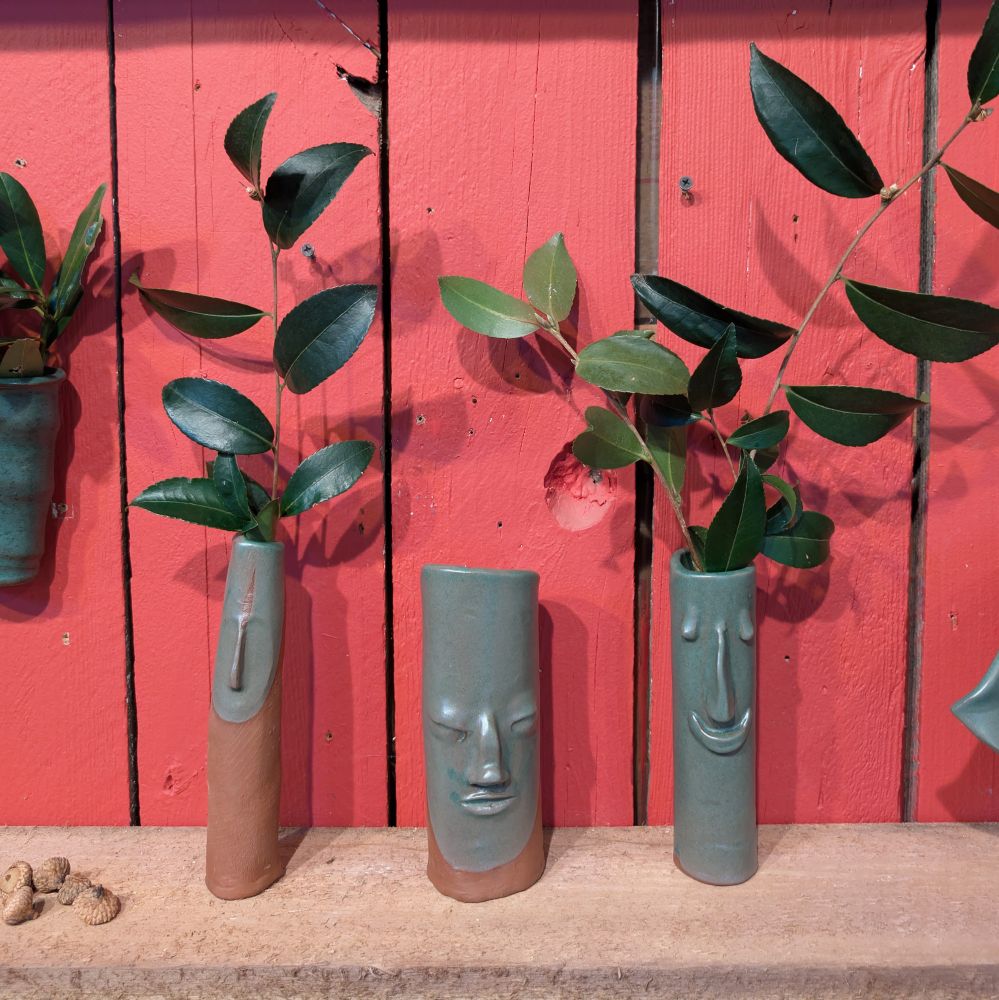 Three green face vases with a red wall in the background. Plants come out of two of them. 