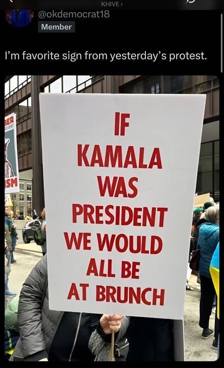 A tweet by @okdemocrat18
 reads, "I'm favorite sign from yesterday's protest." The accompanying photo shows a person at a protest holding a sign that says, "IF KAMALA WAS PRESIDENT WE WOULD ALL BE AT BRUNCH" written in bold red letters. The protest takes place on a city street with other demonstrators in the background, some holding additional signs, including one with a red and black design. The crowd is dressed in winter clothing, suggesting a cold day.
