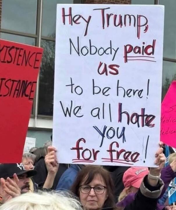 A group of people at a protest holding signs. The main sign reads: "HEY TRUMP, NOBODY PAID US TO BE HERE! WE ALL HATE YOU FOR FREE!" Other signs include one with the word "RESISTANCE" in red and another in pink with partially visible text
