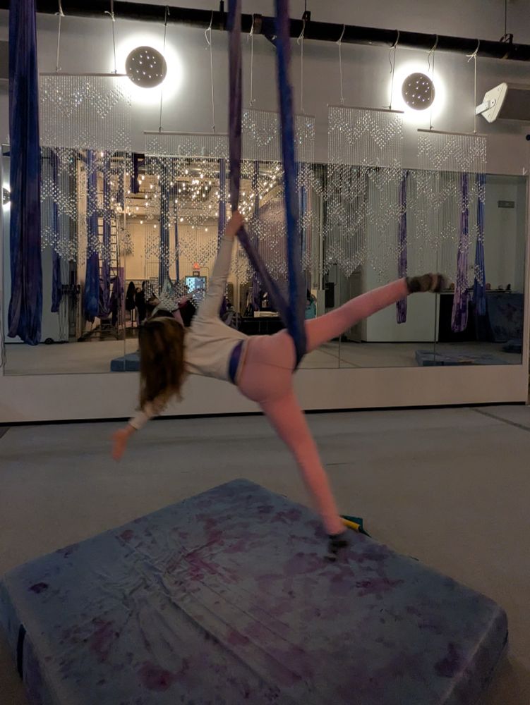Girl in pink pants and I white shirt doing star pose in her aerial swing in front of large mirror