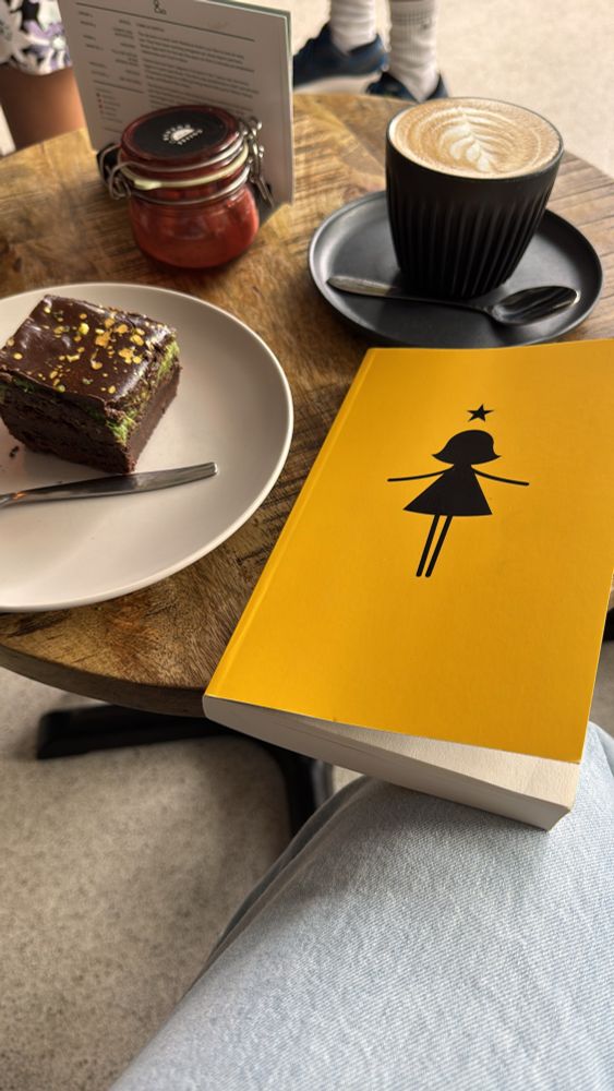 A brownie square, a book with a bright yellow cover and a frothy coffee atop a brown wooden table in a coffee shop. 