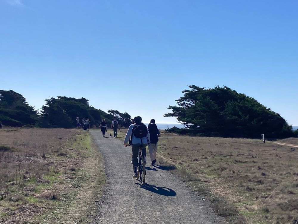People on a trail next to the ocean, walking, on a bike, walking a dog, pushing a stroller. 