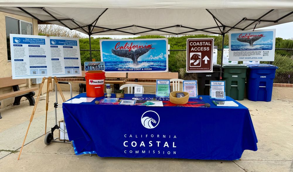 A table with a blue table cloth with the California Coastal Commission logo. Signs are hung from a canopy over the table depicting a Whale Tail License Plate and a Coastal Access road sign, and educational materials are on top of the table.