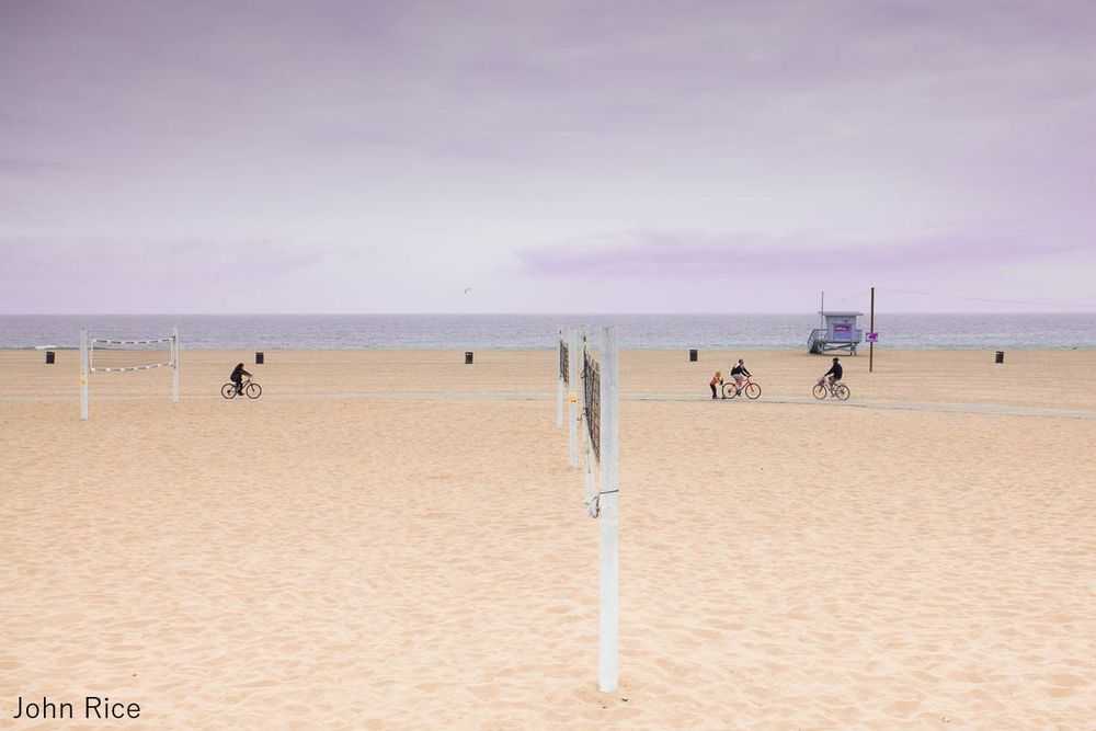 Photo by John Rice shows a broad sandy beach with volley ball nets and a lifeguard stand. A bike path runs through the beach and several people are riding bicycles.