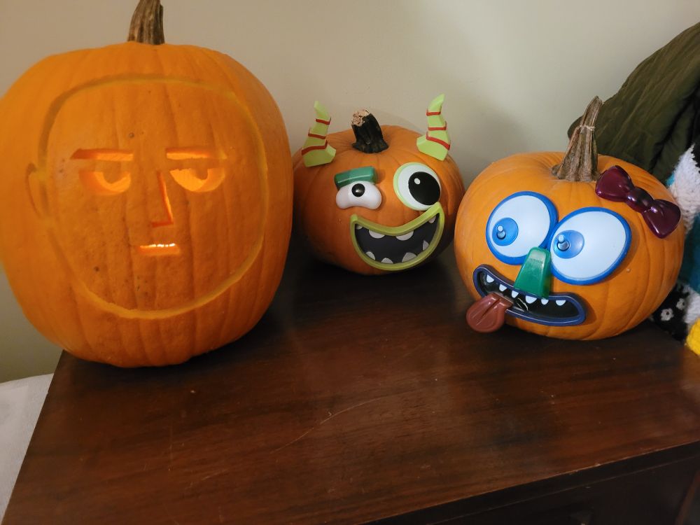 3 pumpkins on a table. One is carved to look like One Punch Man, and the other two have plastic eyes and mouths stuck into the pumpkins giving them wild and crazy faces