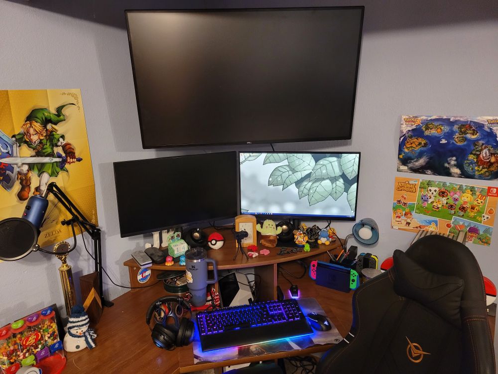 The same desk as from the previous series of pictures, but now with a huge monitor on top. The two monitors beneath are smaller, and the one on the left is dark. There are a lot of gaming peripherals on the desk.