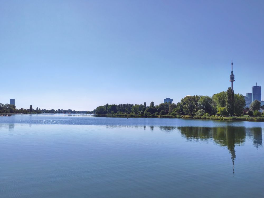 Aussicht flussabwärts über die alte Donau. Kein Wind. Fast spiegelglatte Wasseroberfläche. Der Donauturm spiegelt sich etwas. Ruhe. Am Fahrrad.