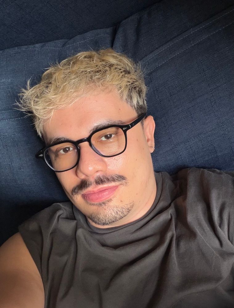 selfie de câmera frontal minha deitado no sofá. o sofá é azul. estou usando um óculos preto e uma regata cinza. sou branco e cabelo tingido de loiro.