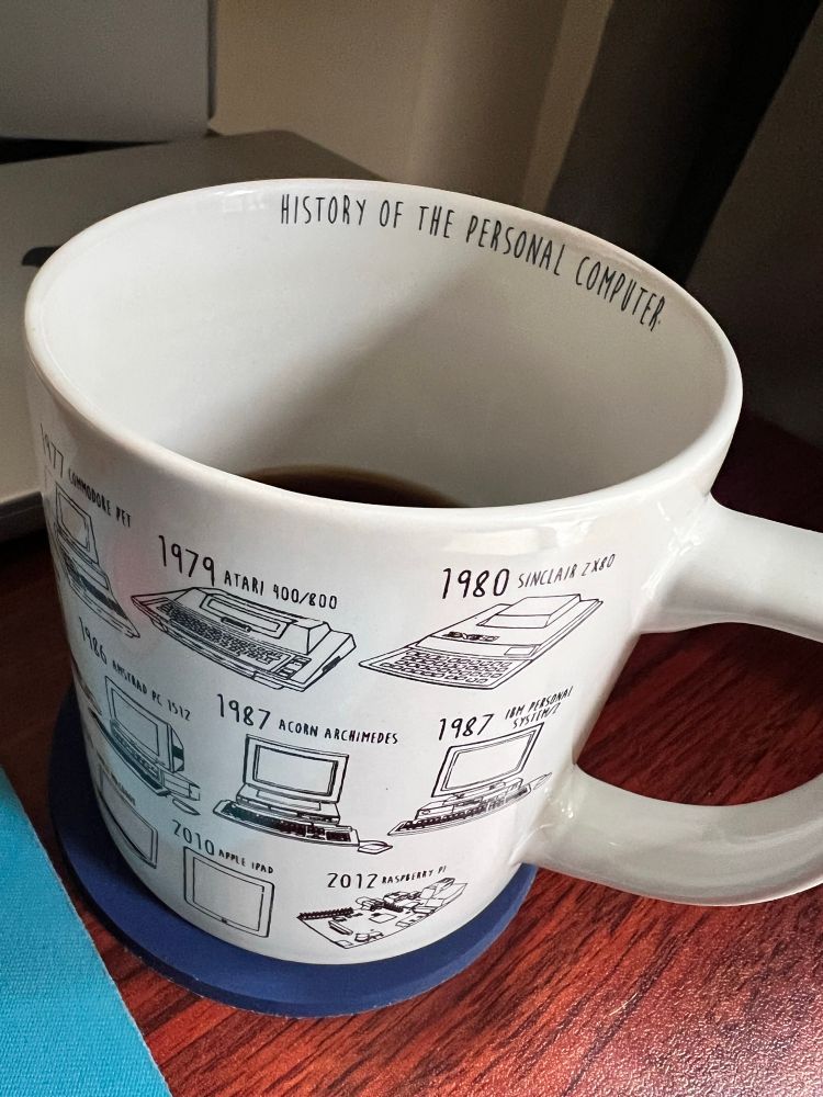A white coffee mug with black coffee. Text on inside wall of the mug says:

History of the personal computer

Three rows of text partially visible on the outside of the mug, depicts computers from 
- 1979 Atari 400/800 ZX80, 1980 Sinclair ZX80
- 1987, Acorn Archimedes, 1987, IBM personal system/Z
- 2010, Apple iPad, 2012 Raspberry Pi

