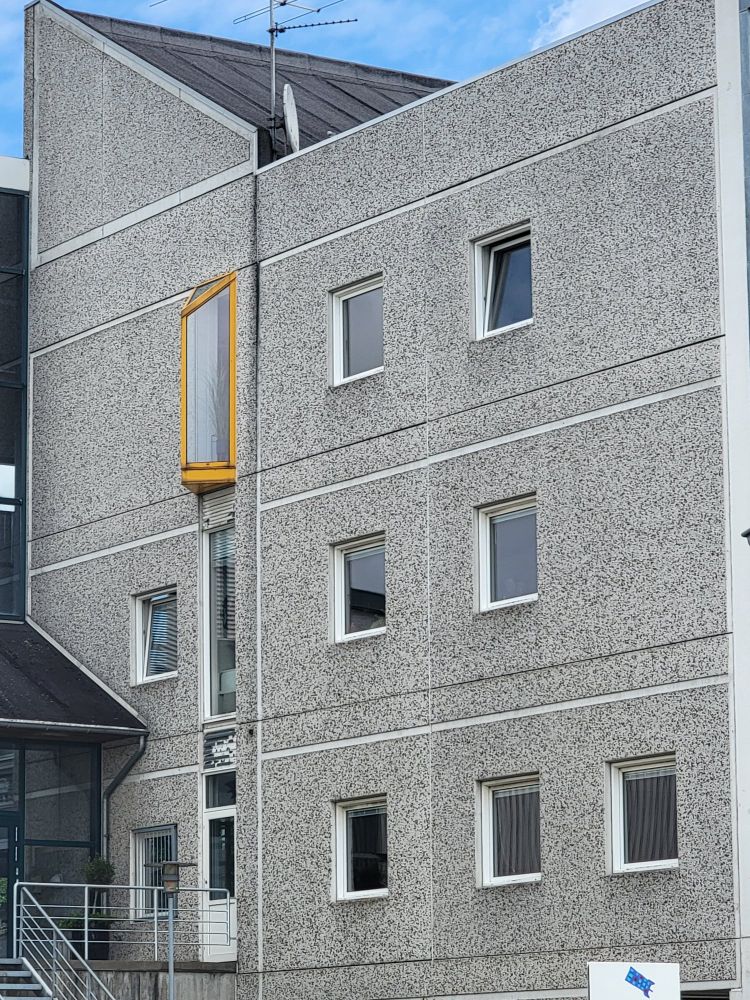 A side of a gray office building in Odense. The only color is the yellow frame around a single window. 