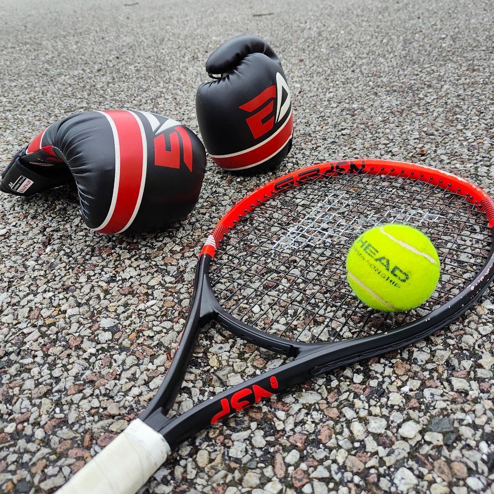 A pair of boxing gloves next to a tennis racket with a tennis ball on it.