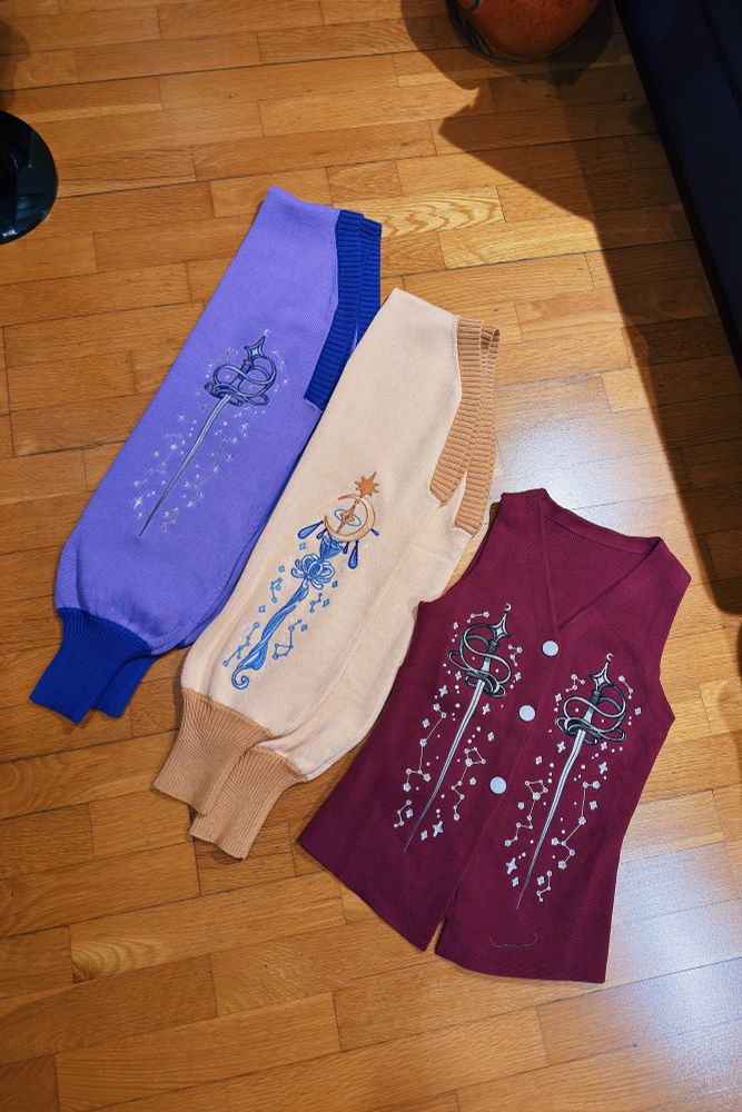 Two knit boleros and sweater vest on wooden floor, with sword and magic staff shaped embroideries. 