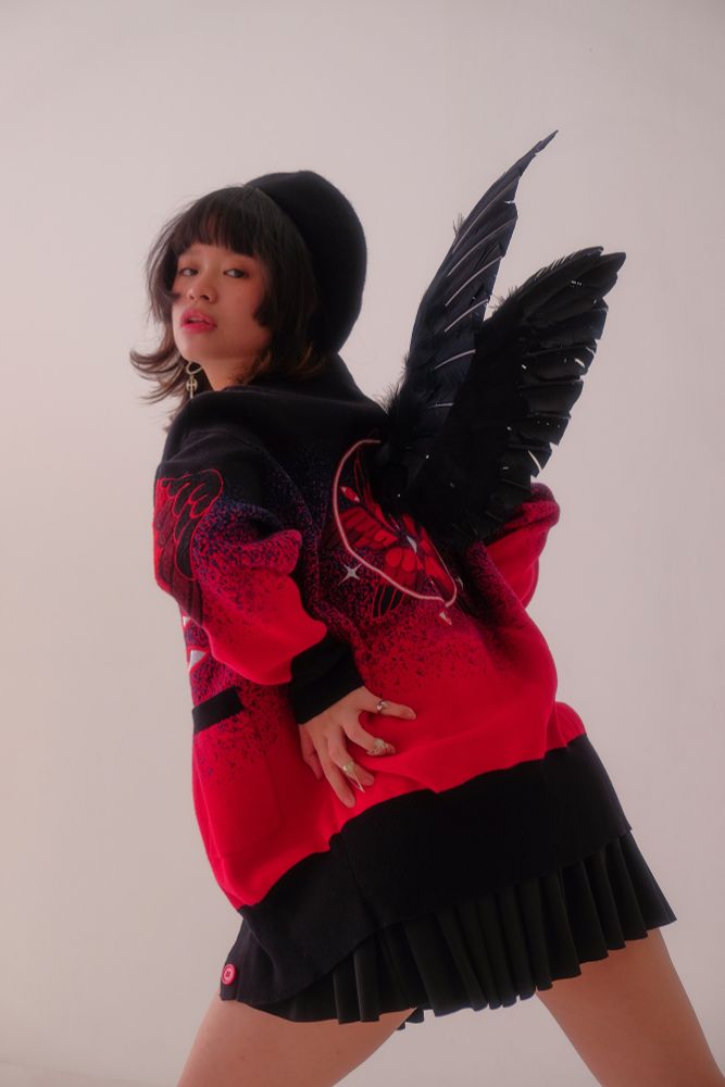 A model wearing black beret, black mini skirt, black angel wings, and black and red cardigan with red eyes, wings, and halo embroideries. 