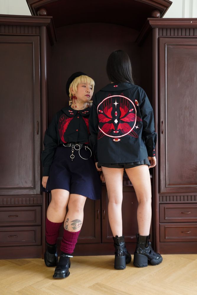 Two models wearing black peter pan collar shirt with red eyes and wings embroideries on the front and collar, and eyes, wings, and halo embroideries on the back. 
