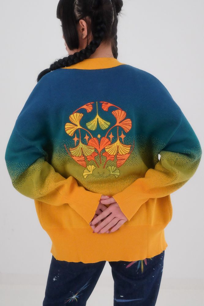 A model facing away from the camera, wearing a blue-yellow gradient knit cardigan with big embroidered ginkgo leaf pattern on the back. 