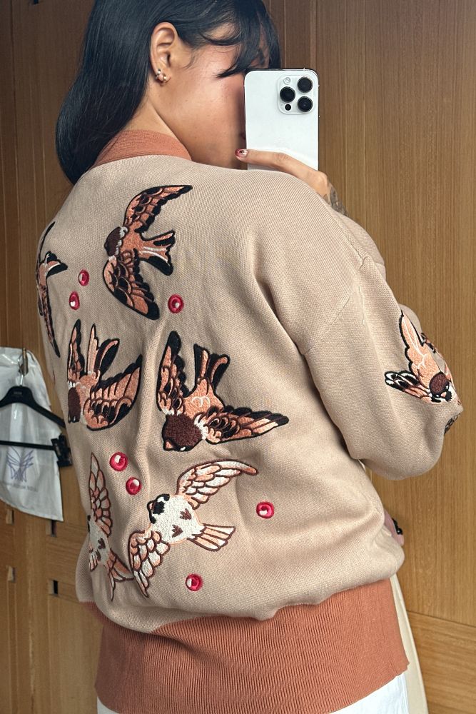 Woman wearing beige cardigan with brown hem and sparrow embroidery on the back.