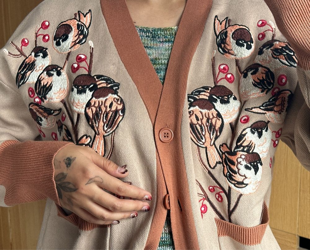 Woman wearing beige cardigan with brown hem and sparrow embroideries on the front.
