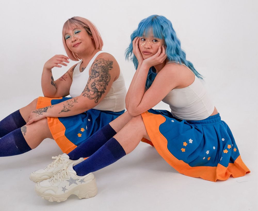 Photograph of two women sitting down, wearing white tank tops and blue and yellow skirts with star embroideries.