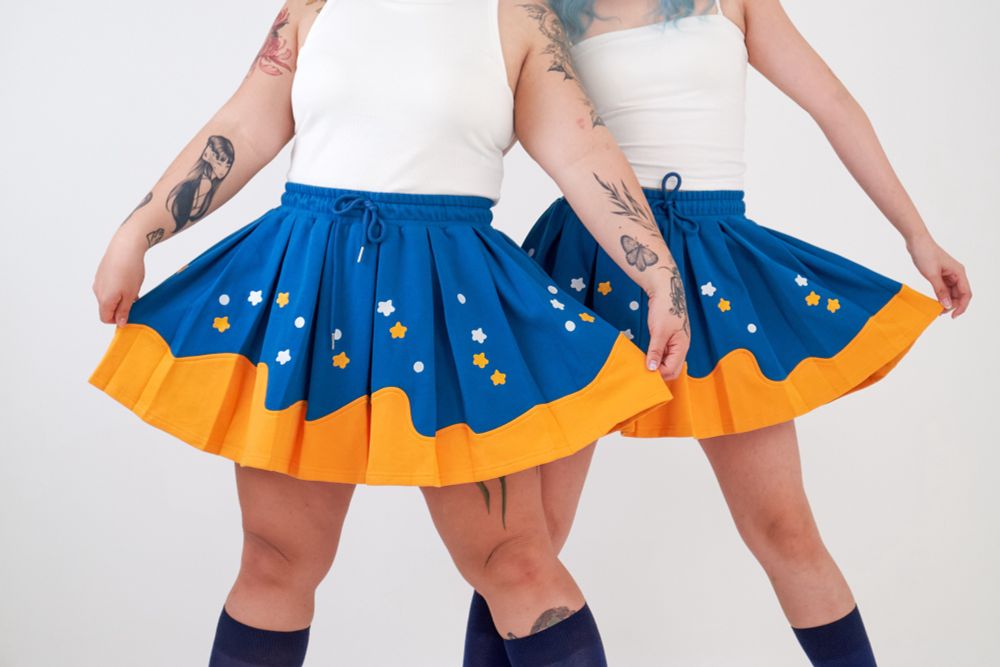 Photograph of two women wearing white tank tops and blue and yellow skirts with star embroideries. 