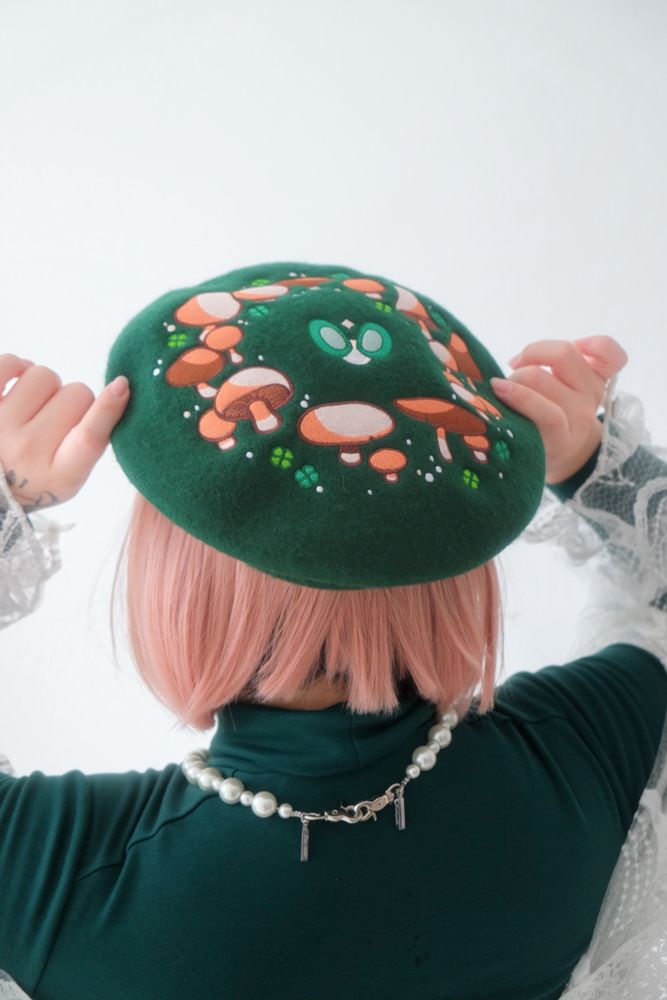A model with pink bob hair modeling a green felt beret with embroidered fairy ring on it. 