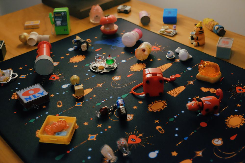Close up of a  desk mat with print of jewels and crows, with tiny trinkets and gachapon toys strewn over it. 