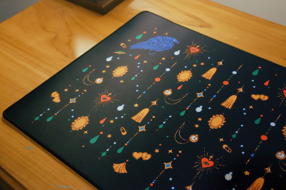 Close up of a black desk mat with jewels, trinkets, and crow print on a wooden desk