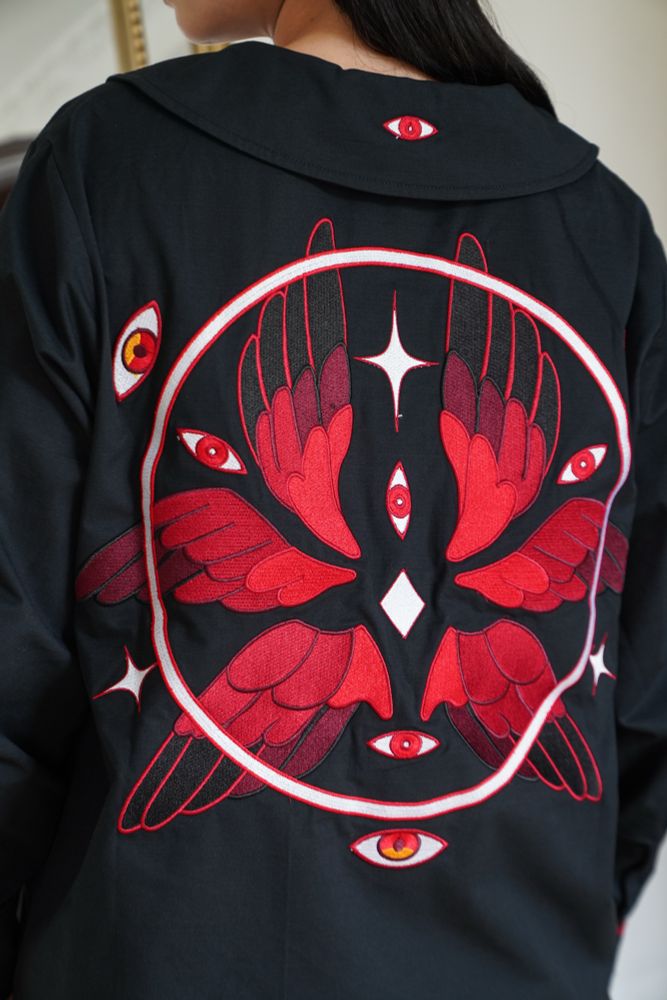 Close up of a model wearing black peter pan collar shirt with red eyes, wings, and halo embroideries on the back. 