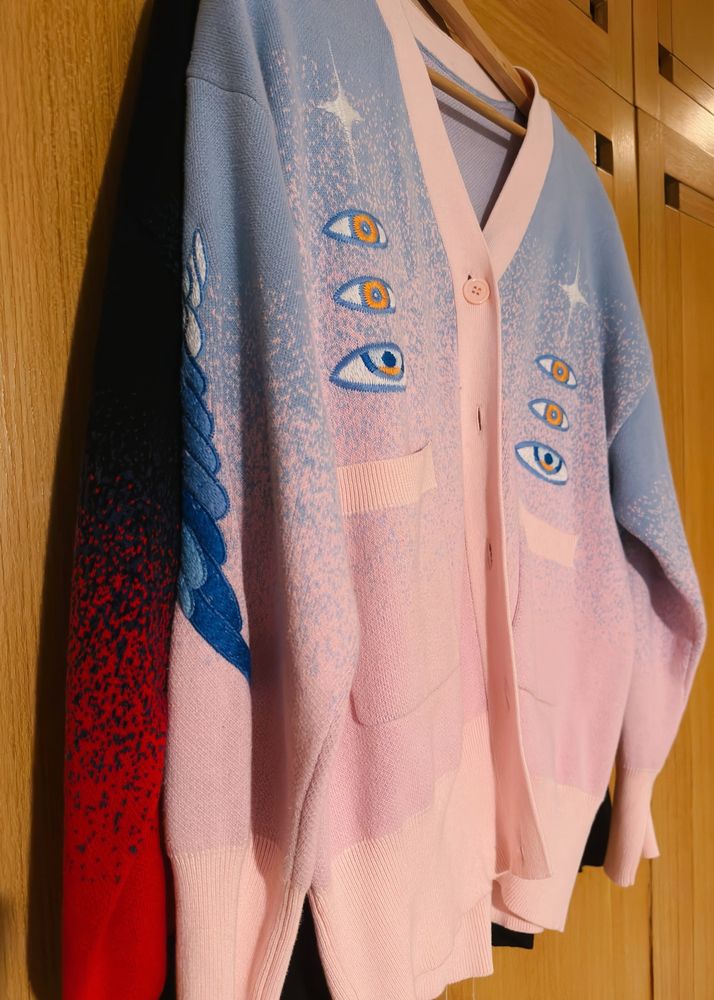Two knit cardigans hanging in front of a wardrobe, one is in black to red gradation and the other is blue to pink gradation, with eyes and wings embroideries. 