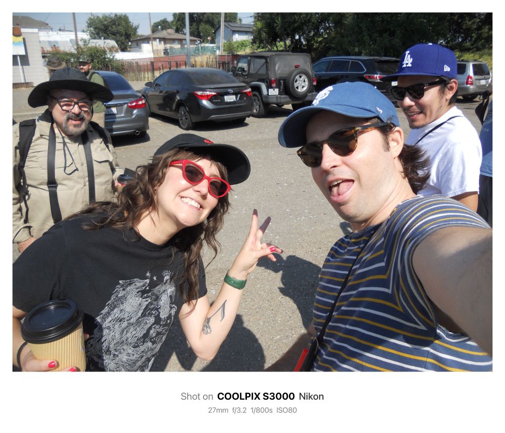 Three people in a parking lot pose for the photo. From left to right is a person wearing a brimmed hat, glasses and khaki shirt with a camera strap and backpack, a person in a black shirt with red sun glasses and a black ball cap holding a coffee to go cup in one hand and making peace fingers with the other hand, a person wearing a blue Giants hat and brown sunglasses sticking out their tongue and holding up the camera used for the photo, last is a person smiling wearing a blue LA dodgers hat and brown sunglasses with a camera strap around their shoulder.

Shot on COOLPIX S3000 Nikon
 27mm f/3.2 1/800s ISO80