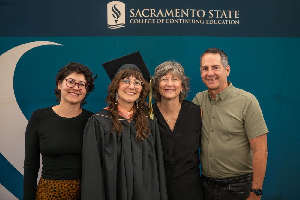 Smiling and posing in front of a green Sacramento State College of Continuing Education backdrop with my parents and younger sister at my MPH graduation.