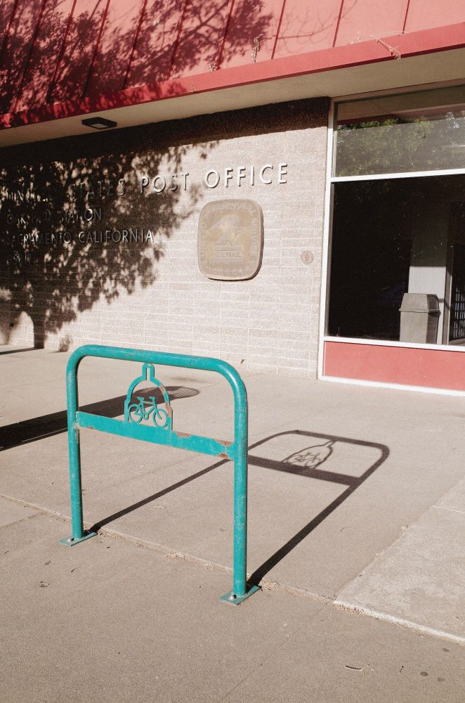 Photo of a U.S. Post Office building in Sacramento, California, bathed in late-afternoon sunlight. In the foreground, a weathered teal bike rack casts a crisp shadow on the concrete, with its cut-out Capitol dome and bicycle motif clearly reflected. Tree branches overhead create dappled shadows on the post office wall, adding texture and depth to the otherwise minimalist scene.