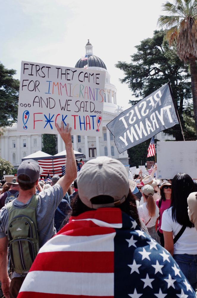 In the foreground a woman in a ball cap  stands with her back to the camera, an American flag draped over her shoulders. She faces the state Capitol building which is partially obscured by a protest sign that reads “first they came for the immigrants…and we said…f*ck that!” And a black flag with white lettering that says “ Resist Tyranny”. 