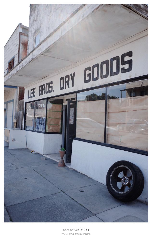 An old white store front with large windows blocked out with brown butcher paper. The white of the building contrasts with a black car tire leaned against the building, black window trim and black block letters “LEE BRUS DRY GOODS” written accross the top.

 Shot on GR RICOH
 28mm f/2.8 1/640s ISO100