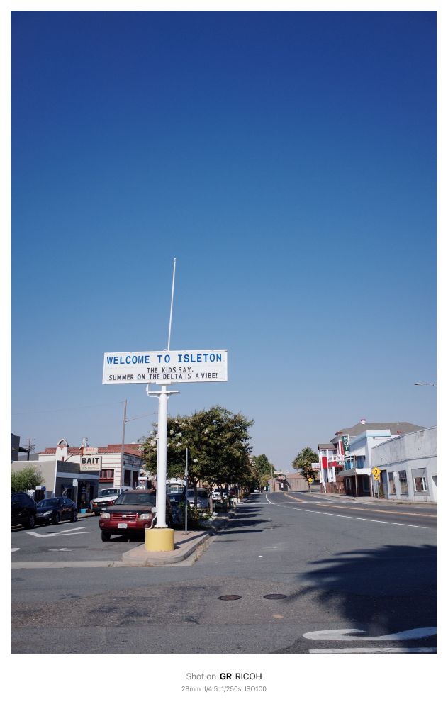 A white sign is highlighted in front of blue sky. The sign reads:
WELCOME TO ISLETON
 THE KIDS SAY.
 SUMMER ON THE DELTA IS A VIBE!
The sign is perched high on a long white pole with roads along both sides. In the background old buildings can be seen lining the roads. One sign says “BAIT” in bold black letters and another says “HOTEL” in vertically stacked letters. On the bottom of the photo a label reads:
 Shot on GR RICOH
 28mm f/4.5 1/250s ISO100